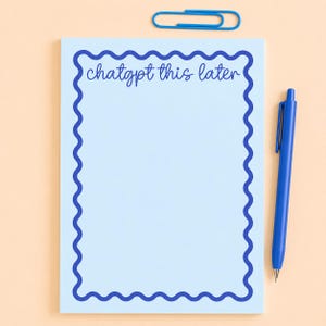 May include: A light blue notepad with a wavy dark blue border and the text "chatgpt this later". A blue pen and paperclip are also present. The notepad is rectangular and sits on a light orange surface.