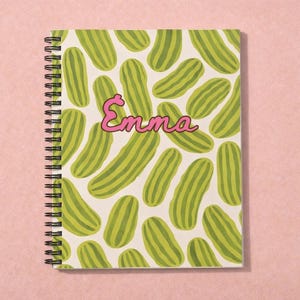 Può includere: Quaderno a spirale con un motivo di illustrazioni di cetrioli verdi su sfondo bianco. Il nome "Emma" è scritto in rosa in corsivo. Il quaderno è appoggiato su una superficie rosa.