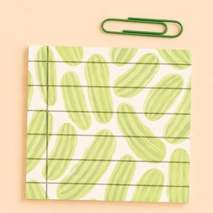 Può includere: Un blocco note quadrato con un motivo ripetuto di cetriolini verde chiaro su sfondo bianco con sottili linee a griglia orizzontali e verticali grigio scuro, accompagnato da una singola graffetta in metallo verde scuro sopra il blocco.
