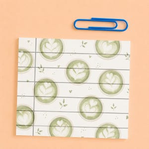 May include: A square notepad sheet featuring a repeating pattern of green matcha latte cups with white heart-shaped foam art, accented by small leaves and dots, crossed by horizontal and vertical gray lines, accompanied by a single blue paper clip.