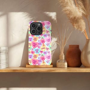 Op de afbeelding: Een telefoonhoesje met een kleurrijk patroon van snoephartjes met verschillende berichten, waaronder "Ik hou van je", "Bel me", "Knuffel me" en "Kus me".