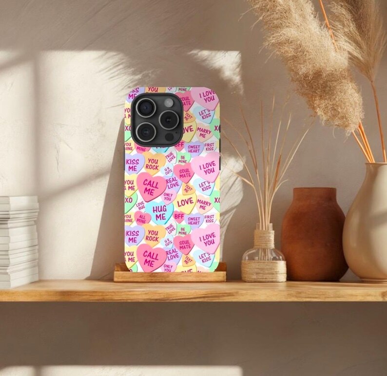Op de afbeelding: Een telefoonhoesje met een kleurrijk patroon van snoephartjes met verschillende berichten, waaronder "Ik hou van je", "Bel me", "Knuffel me" en "Kus me".