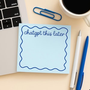 May include: A light blue sticky note with a wavy blue border and the text "chatgpt this later". A blue paperclip, two pens, and a cup of coffee are also on the beige surface, next to a laptop.