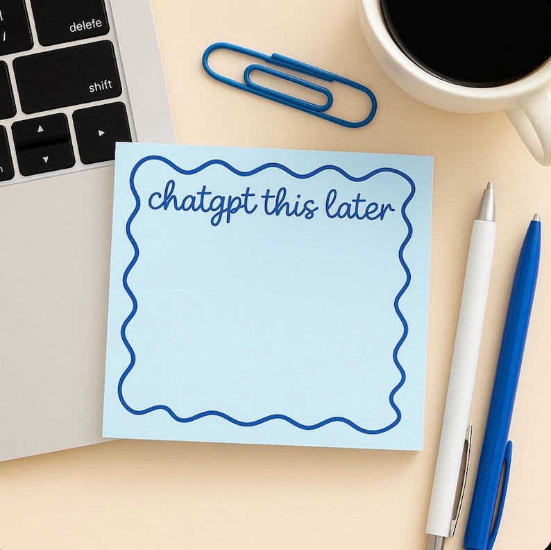 May include: A light blue sticky note with a wavy blue border and the text "chatgpt this later". A blue paperclip, two pens, and a cup of coffee are also on the beige surface, next to a laptop.