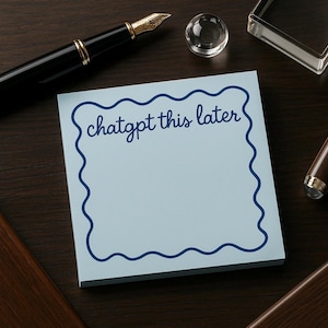 May include: A light blue sticky note pad with a wavy dark blue border and the words "chatgpt this later" in dark blue script. The pad is on a dark wood surface with a black pen and other desk accessories.