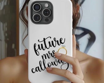 May include: White phone case featuring the text "future mrs calloway" in black script, and a gold ring graphic. The case has a black camera module. The phone case is held up in front of a person.