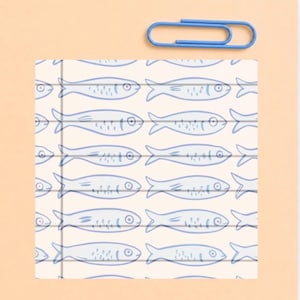 May include: A square notepad with a repeating pattern of light blue line-drawn fish on a cream background, featuring a vertical margin line on the left side. A single light blue metal paperclip is positioned horizontally above the notepad.