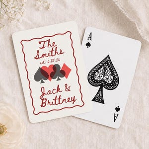 May include: Two playing cards on a white surface. One card features the Ace of Spades. The other card has a red and black design with the names "Jack & Brittney" and "The Smiths" with the date "6.18.26".