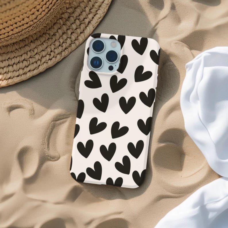 May include: A white phone case with a black heart pattern. The case is lying on a sandy surface.