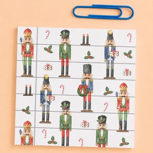 May include: A square notepad featuring a repeating pattern of nutcrackers in red, green, and blue uniforms, interspersed with holly, candy canes, and gift boxes, accompanied by a single large blue paperclip resting above the notepad.