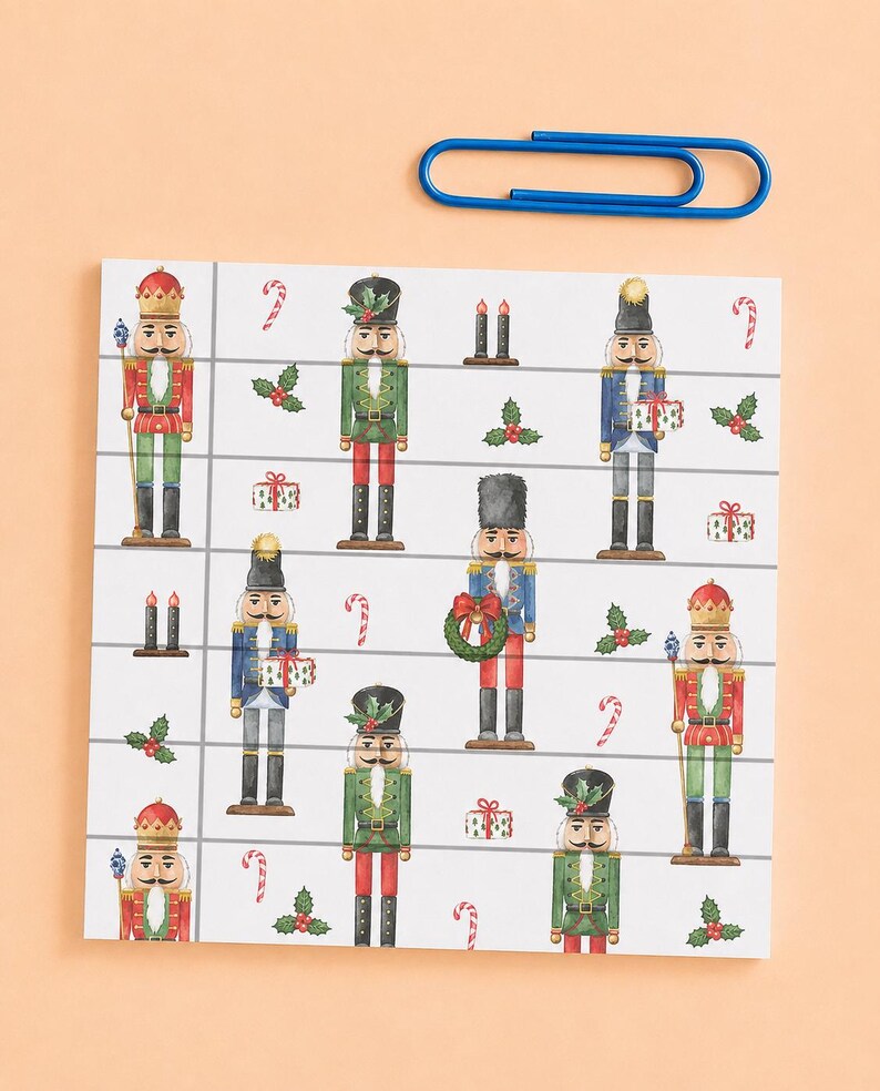 May include: A square notepad featuring a repeating pattern of nutcrackers in red, green, and blue uniforms, interspersed with holly, candy canes, and gift boxes, accompanied by a single large blue paperclip resting above the notepad.