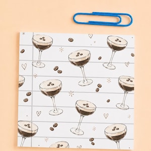 Può includere: Un blocco note quadrato con un motivo ripetuto di bicchieri da espresso martini guarniti con chicchi di caffè, intervallati da piccoli cuori e stelle su uno sfondo bianco con linee grigie orizzontali, accompagnato da una graffetta blu.