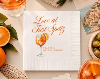 May include: A white paper napkin featuring the orange text Love at First Spritz and Stacey's Bridal Shower, with a watercolor illustration of an orange cocktail in a stemmed glass garnished with an orange slice, centered on the napkin.