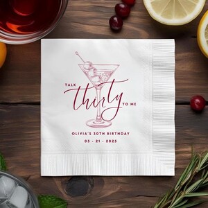 May include: A square white paper napkin featuring a dark red line drawing of a martini glass with olives. The text reads Talk thirty to me, Olivia's 30th Birthday, and 03 21 2025 in a dark red font. The napkin has a textured border on the right side.