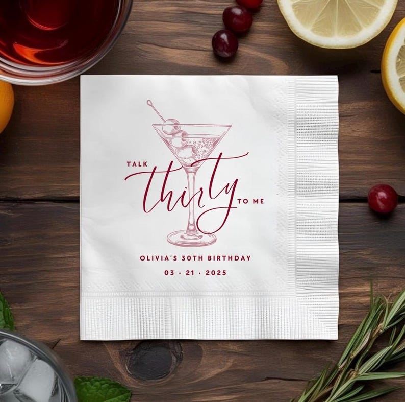 May include: A square white paper napkin featuring a dark red line drawing of a martini glass with olives. The text reads Talk thirty to me, Olivia's 30th Birthday, and 03 21 2025 in a dark red font. The napkin has a textured border on the right side.