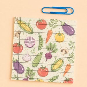 May include: A square notepad with horizontal black lines and a repeating pattern of colorful vegetables including corn, tomatoes, carrots, eggplants, mushrooms, onions, garlic, peas, and zucchini, accompanied by a single blue metal paperclip.