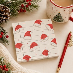 May include: A square notepad with a cream-colored background featuring a repeating pattern of red Santa hats and small red stars. The paper has horizontal lines and a vertical margin line on the left side, printed with a festive holiday theme.