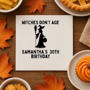 Puede incluir: Una servilleta blanca con el texto "Witches Don't Age" y "Samantha's 30th Birthday" en negro. Una silueta negra de una bruja sosteniendo una escoba y una bolsa también está en la servilleta. La servilleta está rodeada de comida y decoraciones temáticas de otoño.