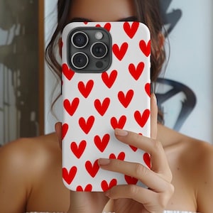 May include: A white phone case with a pattern of red hearts. The case is held up in front of a person's face. The phone case has a camera module with three lenses.