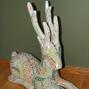 May include: A mosaic deer sculpture with green, orange, and white tiles. The deer is lying down with its head turned to the side. The sculpture is made of small, square tiles that are arranged in a pattern.