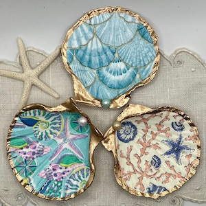 Puede incluir: Tres platos decorativos de conchas marinas con bordes dorados, con diseños pintados de vida marina. Uno tiene un patrón de escamas azules, otro un patrón de estrellas de mar y conchas, y el tercero un patrón de coral y estrellas de mar. Cada plato tiene un acento de perla.