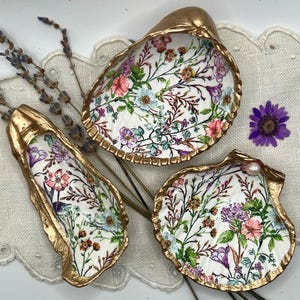 May include: Three decorative seashell dishes with gold rims and floral designs. The shells are painted with colourful flowers and greenery. One shell has a pearl. Dried lavender and a purple flower are also present.