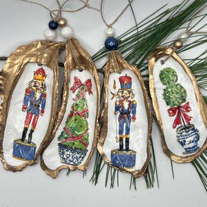 Peut inclure: Quatre décorations de Noël faites à la main à partir de coquilles d'huîtres. Chaque coquille a un bord peint en or et un motif peint sur fond blanc. Les motifs comprennent un casse-noisette, un arbre de Noël et un topiaire. Les décorations sont attachées avec de la ficelle et des perles.