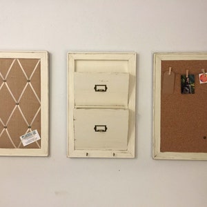 Może przedstawiać: Trzy białe, oprawione organizery ścienne o r&oacute;żnych funkcjach. Najbardziej po lewej stronie znajduje się tablica korkowa z jutowym podkładem i białą wstążką krzyżującą powierzchnię. Organizator środkowy ma dwie szuflady na listy i haczyki poniżej. Najbardziej po prawej stronie znajduje się tablica korkowa z korkową powierzchnią.