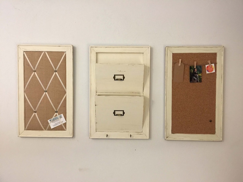 Może przedstawiać: Trzy białe, oprawione organizery ścienne o r&oacute;żnych funkcjach. Najbardziej po lewej stronie znajduje się tablica korkowa z jutowym podkładem i białą wstążką krzyżującą powierzchnię. Organizator środkowy ma dwie szuflady na listy i haczyki poniżej. Najbardziej po prawej stronie znajduje się tablica korkowa z korkową powierzchnią.