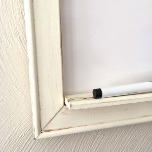 Może przedstawiać: Biała drewniana ramka z białą tablicą do pisania suchą kredą w środku. Biały marker do pisania suchą kredą leży na ramce.