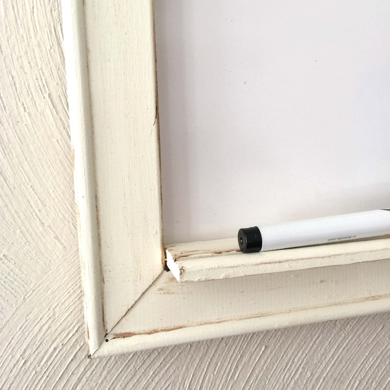 Może przedstawiać: Biała drewniana ramka z białą tablicą do pisania suchą kredą w środku. Biały marker do pisania suchą kredą leży na ramce.