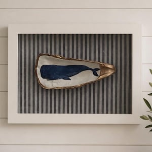 May include: A framed wall art piece featuring a single oyster shell mounted on a grey and white vertical striped background. The interior of the shell is painted with a dark blue whale silhouette and finished with a metallic gold gilded edge.
