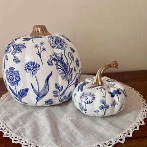 May include: Two decorative pumpkins with a white base and blue botanical illustrations of flowers and dragonflies. The larger pumpkin features a metallic gold stem, while the smaller pumpkin has a curved metallic gold stem.