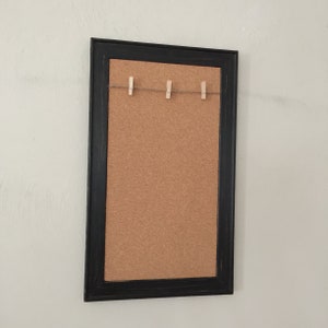 Puede incluir: Un tablero de corcho enmarcado en negro con una cuerda y pinzas para la ropa en la parte superior. El tablero de corcho es una excelente manera de mostrar fotos, notas u otros recuerdos.