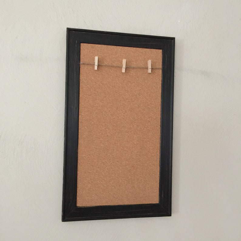 Puede incluir: Un tablero de corcho enmarcado en negro con una cuerda y pinzas para la ropa en la parte superior. El tablero de corcho es una excelente manera de mostrar fotos, notas u otros recuerdos.