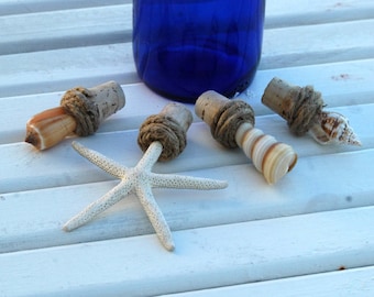 May include: Four bottle stoppers made from cork, rope, and seashells. The stoppers are decorated with a starfish, a seashell, and a spiral seashell. The stoppers are on a white wooden surface.