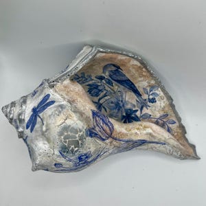 Puede incluir: Una gran concha decorativa con borde y exterior pintados de plata. El interior presenta un diseño floral y de pájaros azul y blanco. Patrones de libélulas y flores azules están pintados en el exterior.