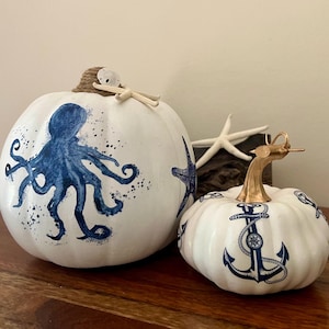 May include: Two white pumpkins with nautical designs. One pumpkin features a blue octopus, the other has blue anchors. A starfish and decorative stem add to the coastal theme. The pumpkins are displayed on a wooden surface.
