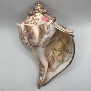 Puede incluir: Una concha marina decorativa grande con borde y detalles pintados en dorado. La concha presenta diseños de coral y conchas marinas en tonos rosa y melocotón. El interior muestra un dólar de arena y otras ilustraciones marinas.