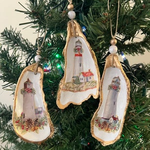 May include: Three handmade Christmas decorations shaped like oyster shells with gold trim. Each decoration features a painted image of a lighthouse with red and white stripes, a red roof, and green foliage.