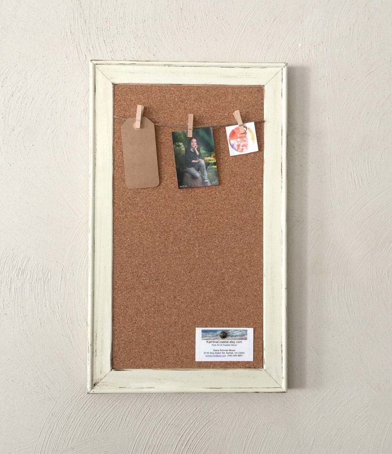 Puede incluir: Un tablero de corcho enmarcado en blanco con una cuerda de pinzas para la ropa que sostiene una etiqueta marr&oacute;n, una foto de una persona y una pintura de acuarela. Una tarjeta de visita est&aacute; fijada en la esquina inferior derecha del tablero de corcho.