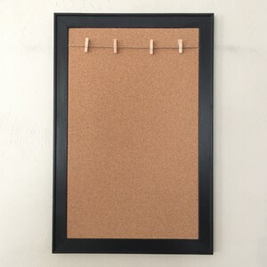 Puede incluir: Un tablero de corcho enmarcado en negro con una cuerda de pinzas para la ropa en la parte superior. El tablero de corcho est&aacute; en blanco y listo para notas o fotos.