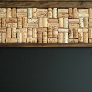 May include: A dark brown wooden frame with a black chalkboard insert. The top of the frame is decorated with a mosaic of wine corks, with various labels and text visible, including "NINE HAT", "ALAMOS", "RADIUS", "LIBCASLIA", "ROMMNIR", and "MAJO".