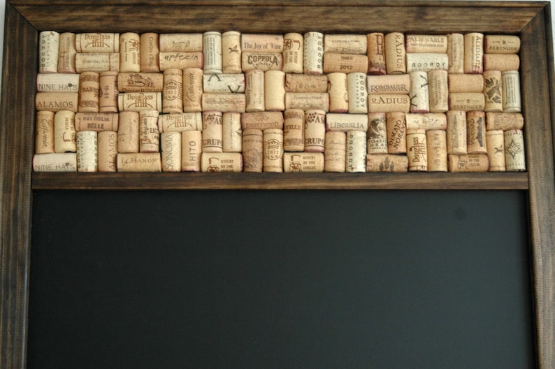 May include: A dark brown wooden frame with a black chalkboard insert. The top of the frame is decorated with a mosaic of wine corks, with various labels and text visible, including "NINE HAT", "ALAMOS", "RADIUS", "LIBCASLIA", "ROMMNIR", and "MAJO".