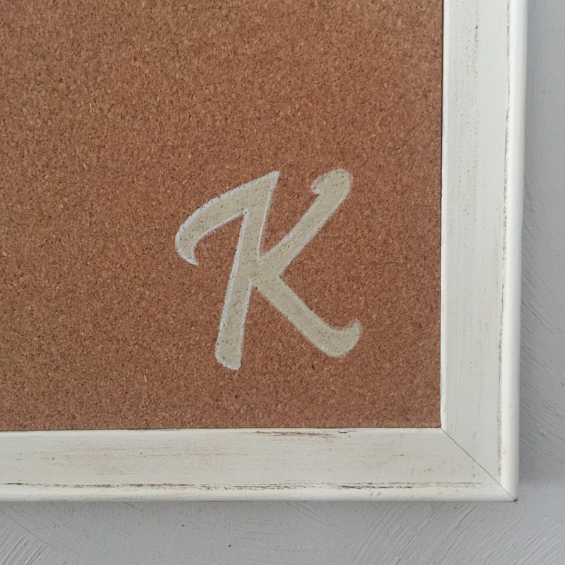 Puede incluir: Un marco de madera blanco con un inserto de corcho. La letra "K" est&aacute; escrita en pintura blanca sobre el corcho.