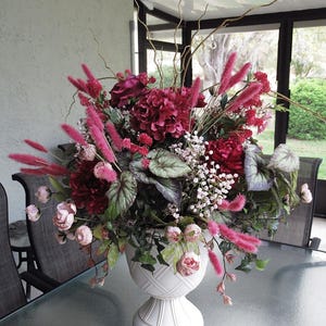 May include: A large floral arrangement in a white urn. The arrangement features deep red hydrangeas, pink feathery plumes, and delicate pink roses. Green foliage and thin, brown branches add texture.