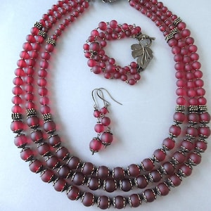 May include: A three-strand red beaded necklace, bracelet, and matching earrings set. The necklace and bracelet feature a leaf-shaped clasp. The beads have a matte finish and are accented with antique silver-toned metal details.