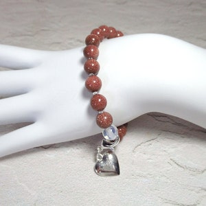 May include: A brown beaded bracelet with a silver heart charm. The beads are made of sandstone and have a speckled appearance.