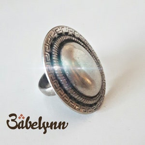 May include: A silver ring with a large, round, domed top. The top of the ring is decorated with a Greek key pattern.