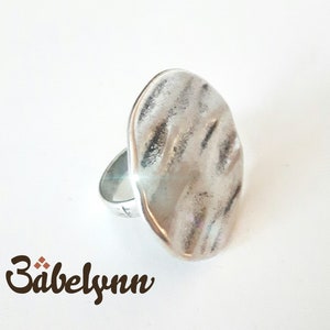May include: A silver ring with a large, textured, oval-shaped top. The ring is made of metal and has a simple band.